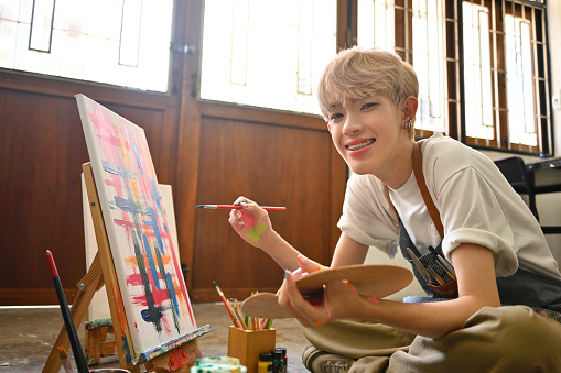 Portrait image of LGBT artist, an Asian male teenager with colored hair, looking at the camera and smiling happily while sitting on the floor and painting.