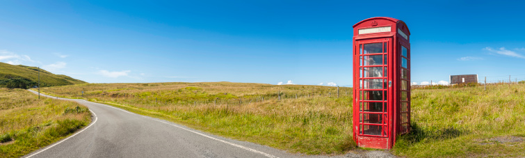 Iconic British red phone box in rural landscape beside country lane and green hills under clear blue panoramic summer skies. ProPhoto RGB color profile for maximum color gamut.