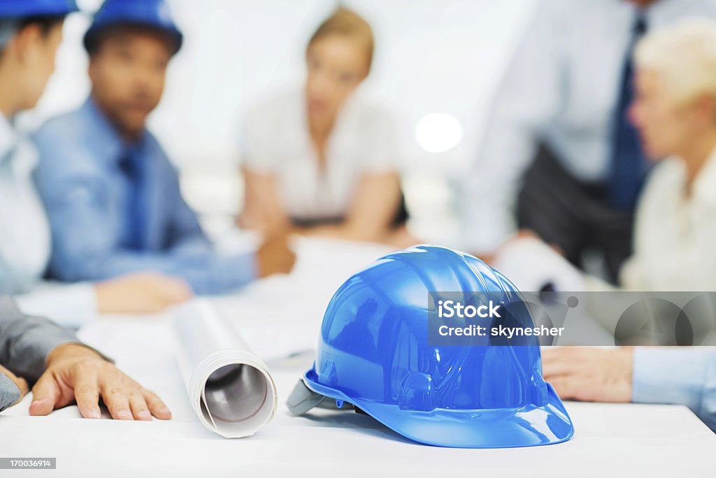Architects on a meeting. Group of successful architects talking about the project in the background. The focus is on the hardhat and rolled blueprints in the foreground.    Adult Stock Photo