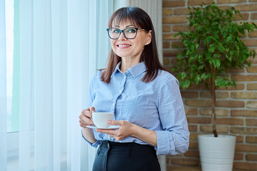 Middle-aged businesswoman with cup of coffee near window, coffee break, smiling female in glasses blue shirt looking at camera. Employees managers work rest