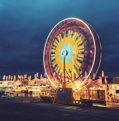 A large ferris wheel spins under twilight skies at a small midway. Long exposure.