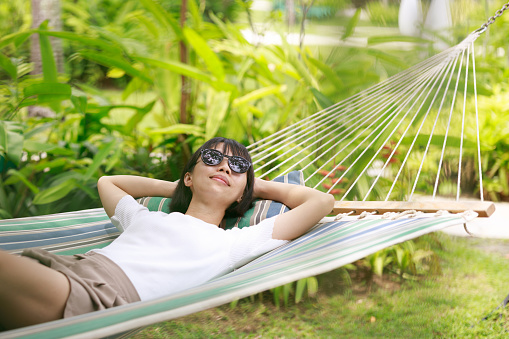 A beautiful Asian woman wearing sunglasses is seen relaxing in a hammock, gracefully enjoying her holiday and spending quality leisure time.