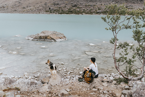 Amidst the serene beauty of a Norwegian landscape, a woman in her twenties takes a breather by a crystal-clear lake, her furry companion contentedly at her side.