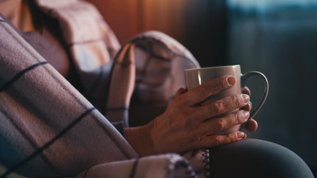 Senior person covered with warm blanket holding cup of tea in hands, close-up