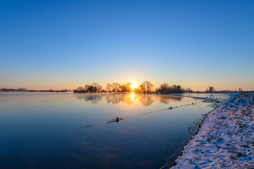 Sunrise over the river IJssel during a foggy cold winter morning in the Netherlands. The water level is high and there is snow and ice on the levees on the shore of the river during this beautiful morning sunrise.