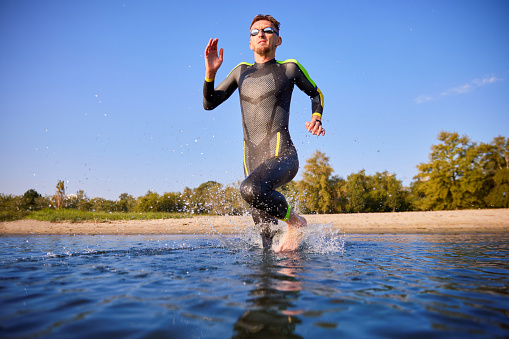 Man, sportsman in wetsuit and goggles training outdoors in morning, running into water, swimming in river. Concept of professional sport, triathlon preparation, competition, athleticism