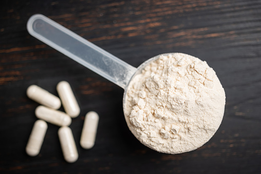 Scoop of whey or soy protein powder, white capsules of amino acids, vitamins and creatine, bodybuilding food supplements, sports nutrition on a dark wooden board, top view.