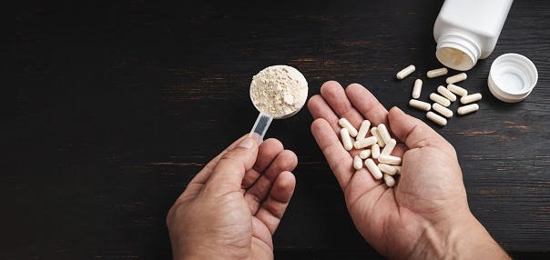 Male hands holds a scoop of whey or soy protein powder and white capsules of amino acids, vitamins, creatine, bodybuilding food supplements, sports nutrition on a dark wooden board.