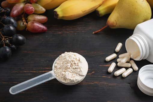 Scoop of whey or soy protein powder, white capsules of amino acids, vitamins, creatine, bodybuilding food supplements and sports nutrition. fruits, banana, pear and grape on a dark wooden board.