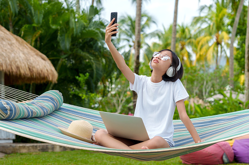 A young Asian teenager girl is seated on a hammock, engrossed in her smartphone as she searches for an internet connection, highlighting the connectivity aspect of modern technology in a relaxed setting.