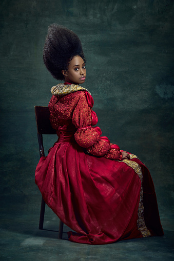 Portrait of beautiful, elegant young african woman, medieval princess in vintage dress sitting, posing against dark green background. Concept of history, beauty and fashion, comparison of eras, ad