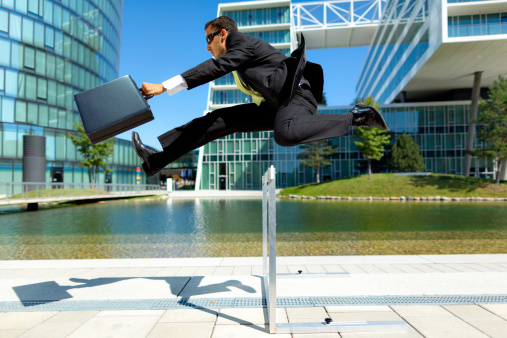 dynamic young business man with briefcase running over hurdle in urban surrounding