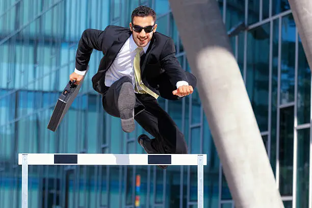 happy young business man with briefcase and sunshades running over hurdle