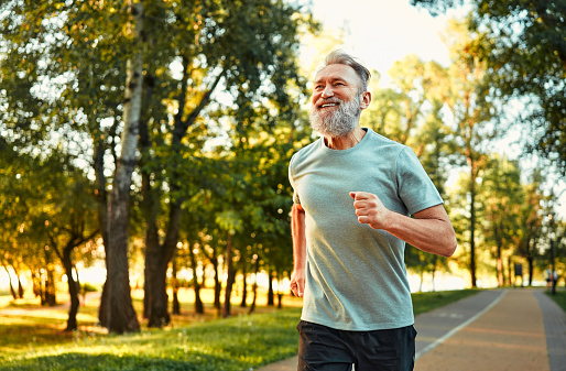 Concept of endurance. Well-shaped senior sportsman feeling joy when running in park. Cheerful male person wearing sport attire strengthening body muscles by every day jogging.