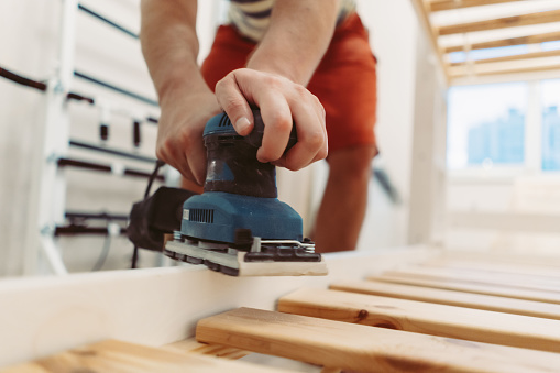 unrecognizable man wearing protective mask rubs wooden child bed with power sander or grinder removing old paint. Image with selective focus