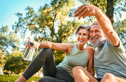 Healthy pensioners after training. Vibrant mature couple in sport outfits taking selfie on smartphone while resting in national park. Encouraged man and woman feeling enjoyment of successful workout.