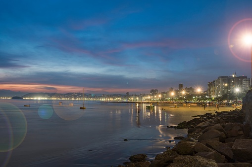 Santos city, Brazil. Santos Beach full of people during sunset. Sea, sand strip and lit-up buildings along the coast.