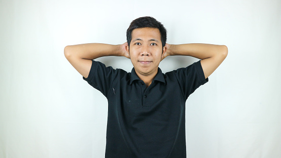 Weekend. Happy asian man wearing polo t shirt relaxing holding hands behind head posing smile on white wall background, lifestyle, emotion expression.