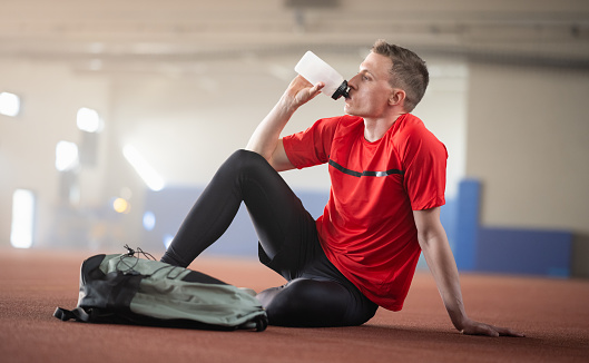 Young male athlete sitting on the floor, drinking water and resting after hard training. Sport and healthy lifestyle concept.