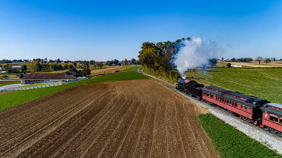 A Drone View From Behind and Above of a Steam Passenger Train Traveling Thru Farmlands and Blowing Smoke on a Sunny Fall Day