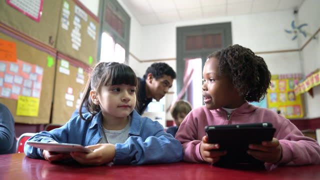 Happy diverse little girls having fun during IT class using tablets while talking