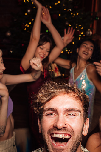 Group of friends celebrating New Year together at a party in the North East of England. They are having fun dressed fashionably dancing together. Focus is on one man who is close to the camera laughing while looking at it.