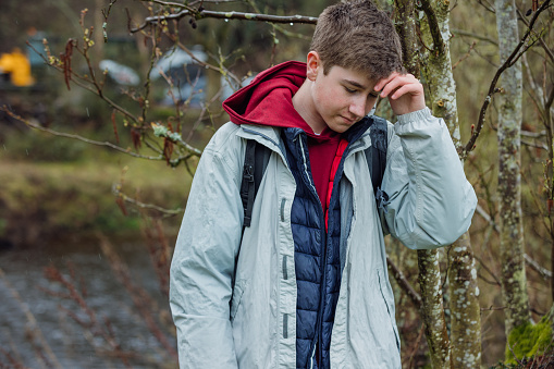 A medium shot of a young male student wearing outdoor clothing and a backpack on an overcast day in Northumberland. He is on a school field trip with his peers and teachers. He is standing alone in the woods with his hand on his head looking sad.