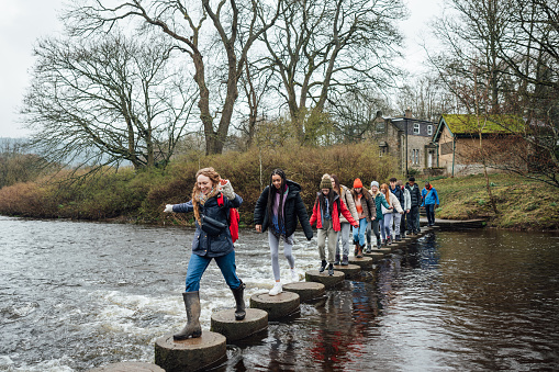 A wide shot of a large group of students and their two teachers wearing warm outdoor clothing on an overcast day in Northumberland. They are on a field trip and are crossing a river by walking over stepping stones, their female teacher is leading the group.

Video also available of this scenario