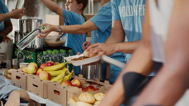 Poor Wheelchair User Receiving Free Food At food drive, poor female wheelchair user receives free food and provisions. Multiethnic volunteers in blue t-shirt distributing fresh fruits and hot meals to less privileged. Close-up, tripod. donation box stock pictures, royalty-free photos & images