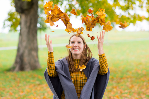 Beautiful young woman in fall clothing, a grey shawl and mustard yellow plaid turtle neck shirt, is laughing as she is throwing fallen autumn leaves into the air celebrating the changing seasons in a park outdoors.