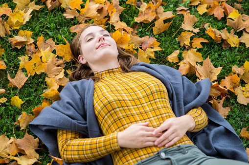 Beautiful young woman with brown hair wearing fall style clothing, yellow plaid and a grey shawl, is laying down with her eyes closed in the fallen Autumn leaves in a park outdoors.