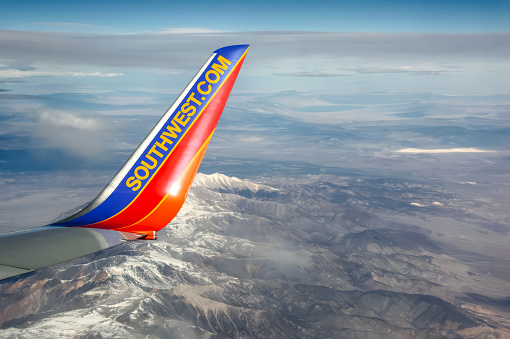 A wing tip of Southwest Airlines Boeing 737 flying over snow-capped mountains. The blue and orange colors contrast with snowy peaks and clear sky. The image captures Southwest's spirit of adventure.