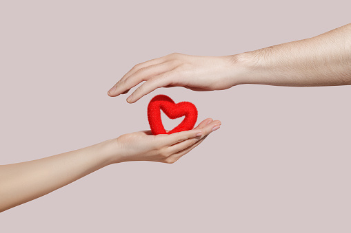 Red heart in woman and man hands on pink background. Hope and family concept, world heart day, world health day. Copy space for advertisers.