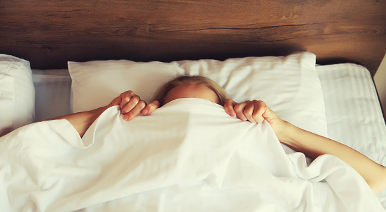 Healthy sleep, woman lying in bed under white blanket or duvet hiding her face in bedroom at home