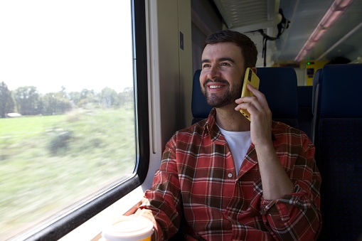 A handsome man in his forties is sitting on train while making a call. He is very handsome and has a nice facial hair. He smiles and looks away during his relaxing trip.