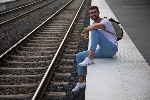 Image where a cheerful lonely looking man is waiting at a train station. He sits down on the platform after his train already left.