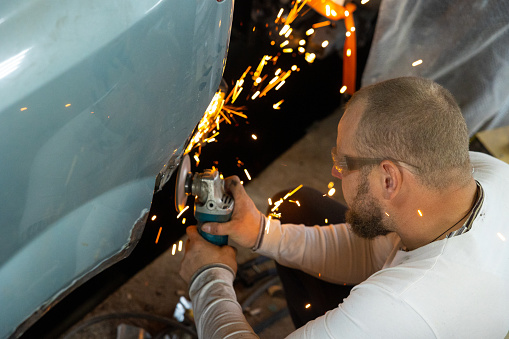 The mechanic uses a grinding machine to remove rusty metal from the surface of a car. The professional auto mechanic repairs the car in his workshop.