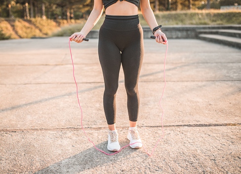A young unrecognizable athletic woman dressed in sportswear is training outdoors and jumping with skipping rope