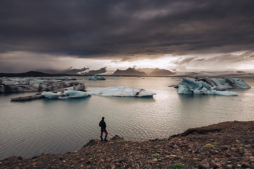 A man is watching icebergs in the ocean in the wild nature of Iceland. There is a stormy dramatic sky in the background. A majestic view of wild, dramatic, and stormy nature in Iceland.