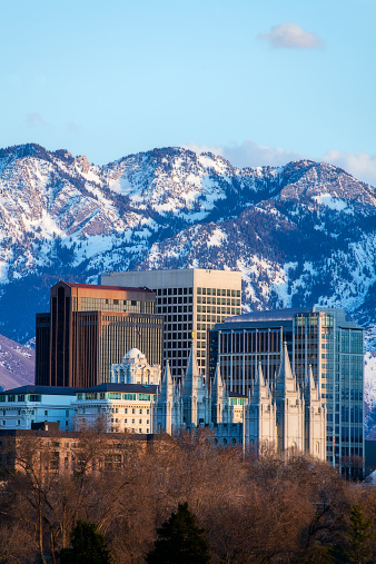 A royalty free DSLR vertically oriented photo of the skyline, with copy space, of Salt Lake City, Utah, USA in early spring at sunset with alabaster white, gray, and amber buildings in the foreground; and gray, snowcapped mountains and blue sky in the background. Includes: Salt Lake temple of the Church of Jesus Christ of Latter-day Saints (the Mormons), Joseph Smith Building), Eagle Gate Building, bank high rise buildings, and apartment condominiums of City Creek Plaza.