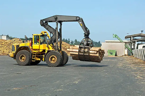 Wheel loader loading timber in saw mill. Grapple full of timber near barking line.