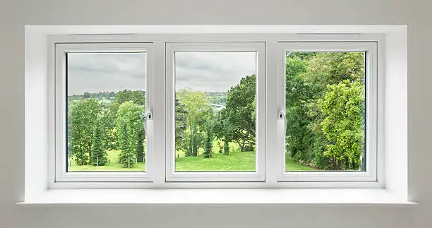a set of three windows in a white frame surrounded by a light grey wall with views over a lovely countryside garden. with mature trees.