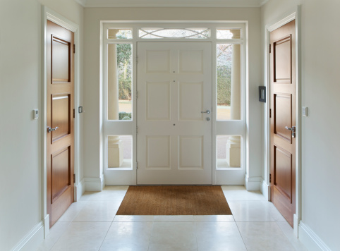 a front door entrance to a grand house. The wood panelled front door and frame are painted white whilst either side, also in white frames are two wooden doors for two separate cloakrooms. Inset into the cream coloured marble floor is bristle floor mat. Through the glass that surrouds the front door frame one can see part of a carriage drive whilst two columns sit either side on the outside of the front door.