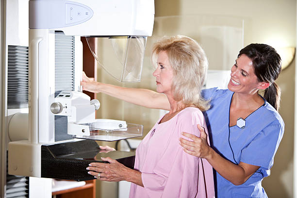 Nurse with patient getting mammogram A woman in her 50s getting a mammogram.  She is standing at the machine wearing a pink gown.  A female technician is atanding behind her, smiling, in blue scrubs, with one hand on the patient's shoulder and smiling. x ray image medical occupation technician nurse stock pictures, royalty-free photos & images