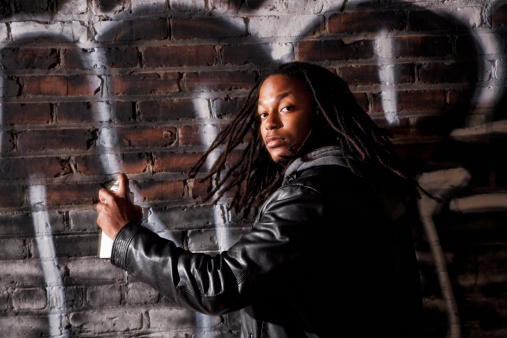 Young black man (20s) holding paint can in front of brick wall covered with grafitti.