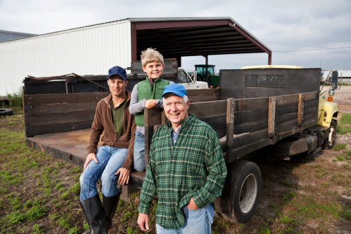 Three generations of men on the back of a truck on the family farm.  The grandfather, a senior man in his 60s, is the focus of the image, standing with his hand in his pocket, smiling at the camera.  His adult son and young grandson are behind him on the truck.  They are wearing jeans, work boots and jackets or flannel shirt.  Everything is brown or green, earthy colors.