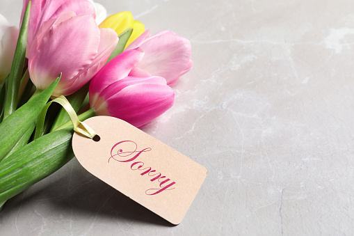 Word Sorry written on label and bouquet of tulips on light grey marble table, closeup. Space for text