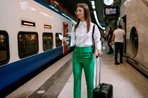 A young woman stands stationary on the train station platform, suitcase by her side, as she scrolls through her phone