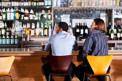 Rear view of multi-ethnic men having drinks at bar, watching TV screen off to the side.