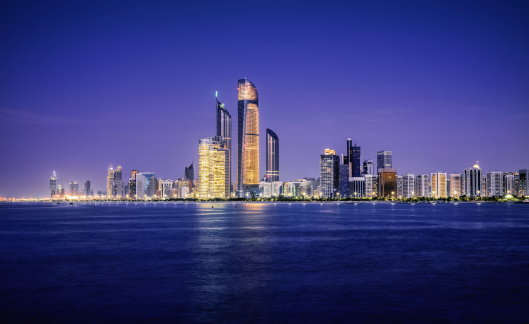 The skyline of Abu Dhabi at sunset, taken from the distance over water.  All of the buildings have their lights on, and the large towers in the middle are the Central Market Towers.  The sea is very tranquil, and the fading light lingers behind the city.